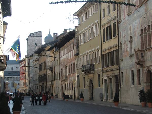 People walk along Trento's Via Belenzani past frescoed buildings and Christmas decorations