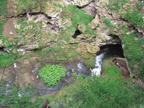Waterfall over limestone rock with cave, green vegetation and tourists on the right
