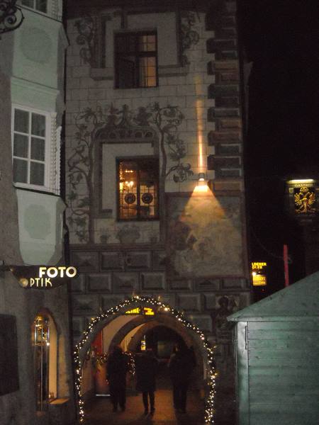 Christmas-lit archway under a frescoed building, Foto Ottik sign, people passing at night