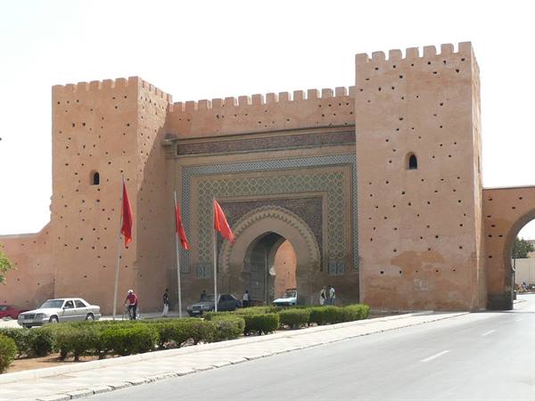 Monumental earthen gate with zellige tiles, red flags and pedestrians in Meknes