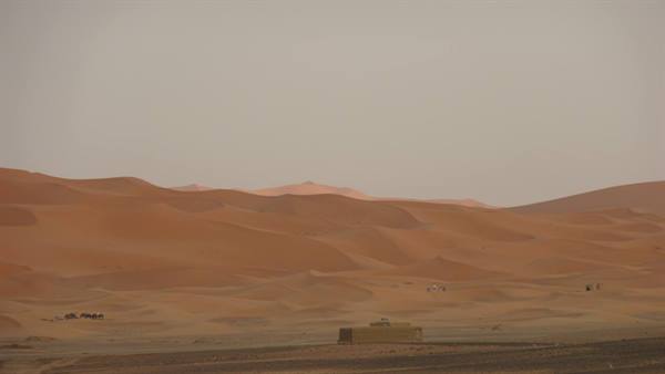 Orange sand dunes stretch across the desert, a concrete building in the foreground, hazy sky