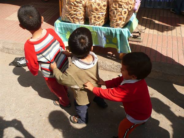 Three children in a line on a sidewalk, near a stall of bagel-like breads