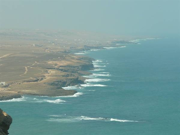 Arid cliffs plunging into turquoise sea, breaking waves, barren land under hazy sky