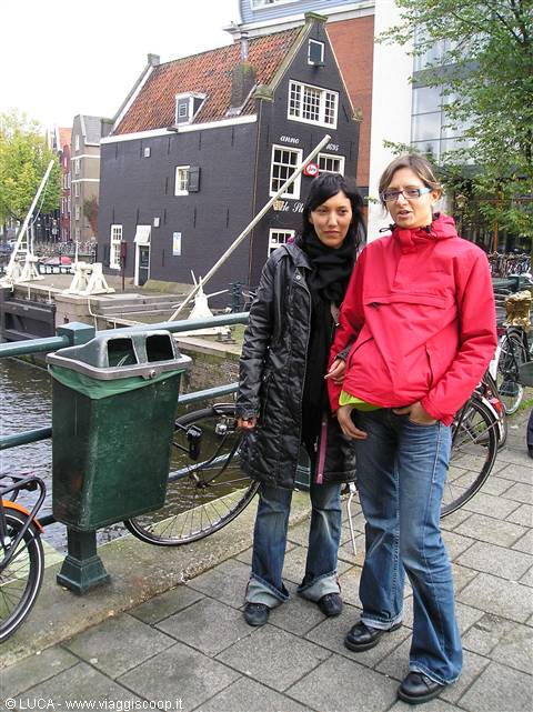 Two tourists in jackets and jeans beside a black wooden house on a canal