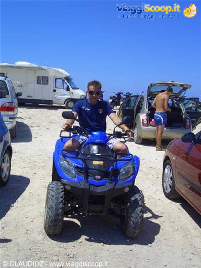 Smiling man riding blue ATV on sandy lot, cars and camper nearby