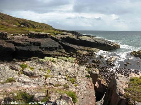 Reddish rocky cliffs meet the sea, green grass and cloudy sky, Scotland