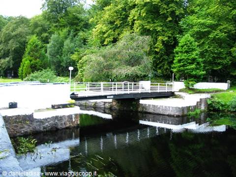 White metal bridge over a canal lock, green trees reflected in dark water