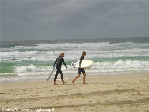 Two surfers in wetsuits walk along the beach carrying their boards