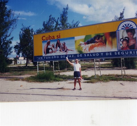 Man poses under a yellow Cuba Sí billboard on sand and trees