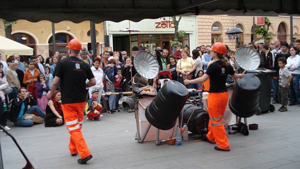 Performers in orange helmets drum on metal barrels for a crowd in the square