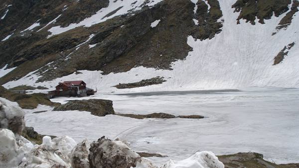 Wooden mountain hut by frozen lake, snow and rocks around