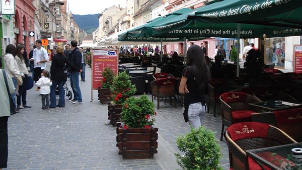People strolling past cafes with green umbrellas and wooden flower planters, Brașov