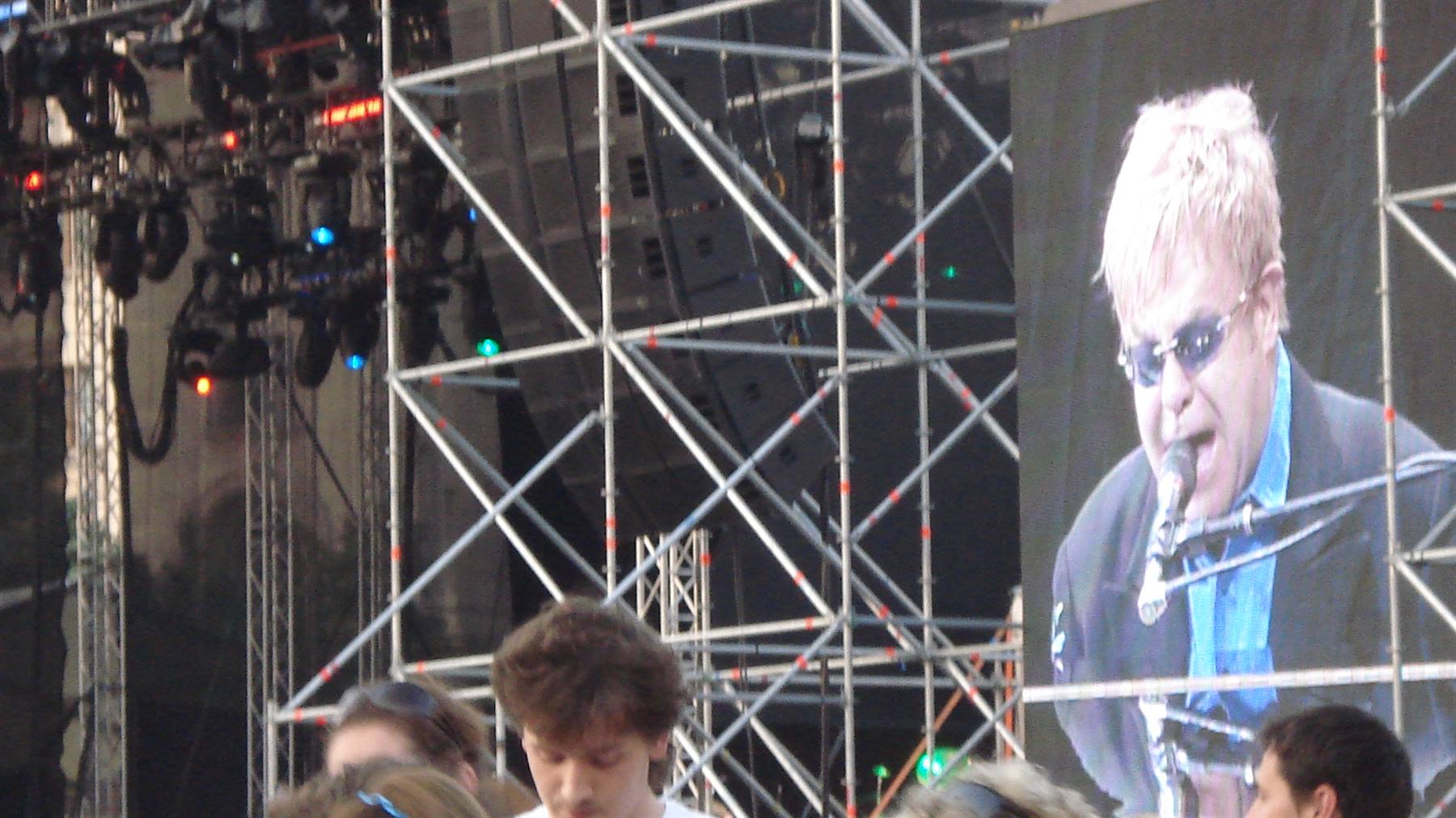 Giant screen shows Elton John singing amid metal scaffolding and concert crowd
