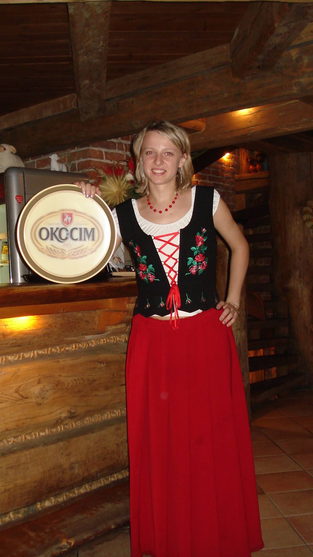 Waitress in traditional costume with red skirt beside an Okocim barrel in a wooden tavern