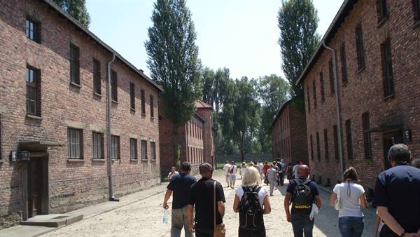Visitors walk between Auschwitz I brick barracks on gravel under sun