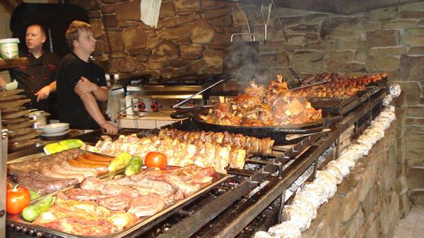 Cooks behind a huge grill full of meat, sausages and vegetables, stone walls