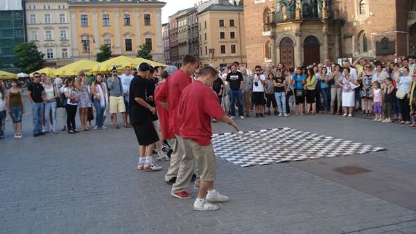 Breakdancer in red shirt dancing on checkered mat in square, crowd watching