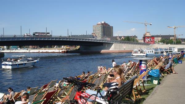 People relaxing on wooden deck chairs by the Spree river in Berlin, bridge and boats