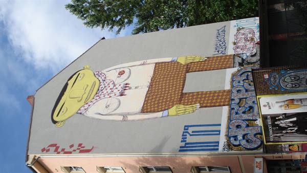 Giant mural on building facade: yellow figure with checkered trousers, graffiti and posters