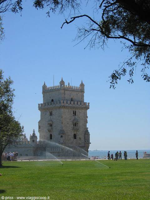 Belem Tower in stone stone beside lawns and river, tourists strolling by