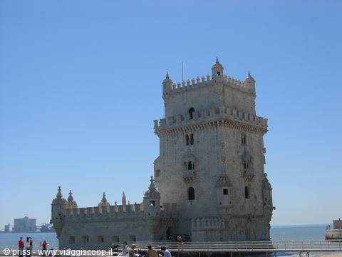 Tourists walk past the stone Belém Tower under a clear blue sky