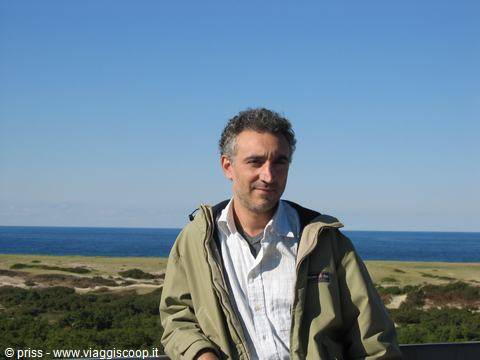 smiling man in green jacket in front of dunes and blue sea under clear sky