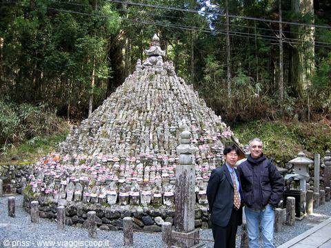 Two tourists beside a pyramid of stone Jizo statues at a forested temple
