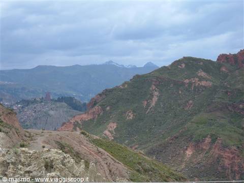 Green and red rock valleys under grey clouds, snowy peaks in the distance