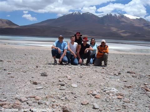 Group of tourists posing on rocky ground in front of lagoon and snowy mountains