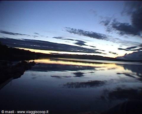 Calm lake water mirrors clouds at dusk, dark hills on the horizon