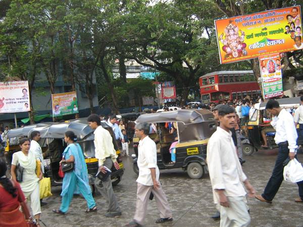 Crowd crossing street among auto rickshaws and a double-decker bus, colorful billboards on trees