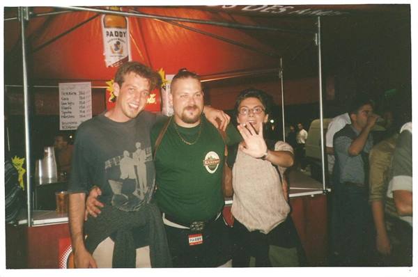 Three smiling friends at the Paddy bar stall, red tent lit at night
