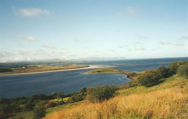 Blue bay with sandy beach and grassy hills under a partly cloudy sky