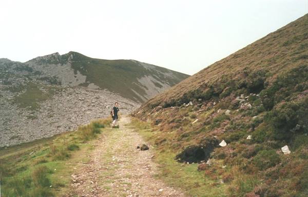Hiker walking on grassy trail between bracken-covered hills under grey sky