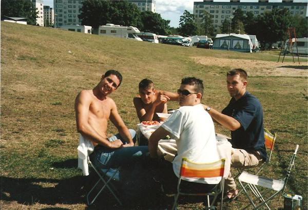Four friends eating on folding chairs on the grass, campers and buildings behind