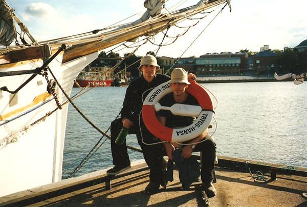 Two people holding a Trigge-Hansa lifebuoy on a dock beside a tall ship