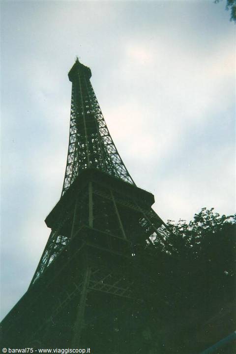 The Eiffel Tower in wrought iron seen from below among trees and cloudy sky