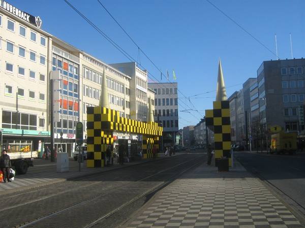 Yellow and black checkered arches with spires cross tram tracks in a city