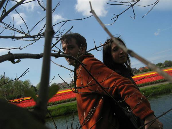 Two people among bare branches in front of red tulip fields and a canal