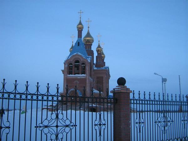 Red Orthodox church with golden domes behind wrought iron fence, snow at dusk