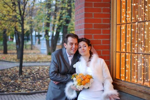 Bride and groom by lit window, white fur coat and orange bouquet, autumn park behind