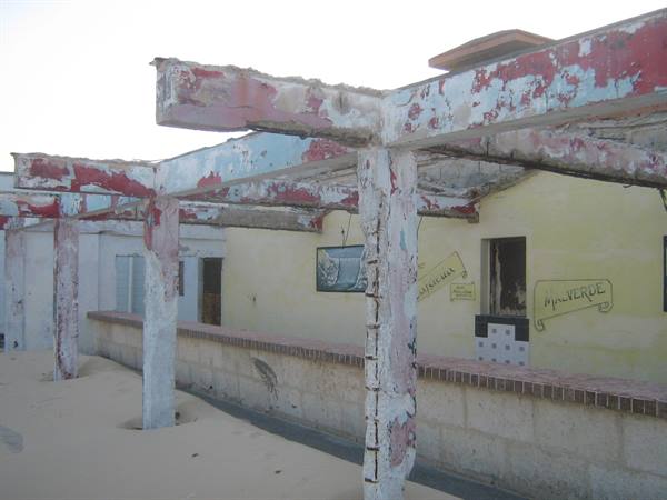 Weathered red and blue painted beams above yellow walls half buried in sand