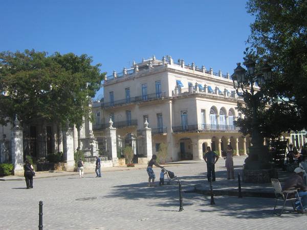 People stroll past a white colonial palace on a sunny paved square, Havana
