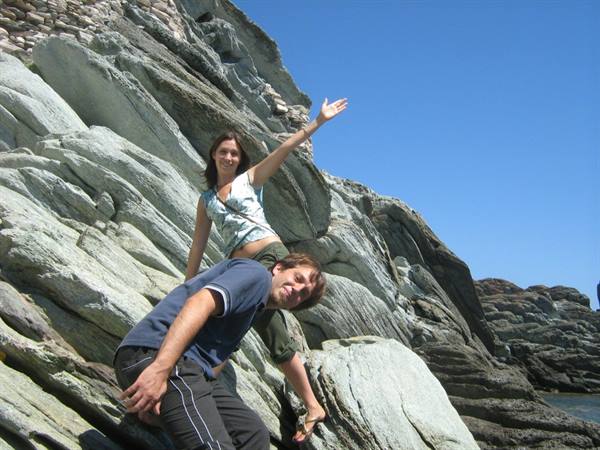 Smiling couple on slippery gray rocks, she waves with raised arm, blue sky