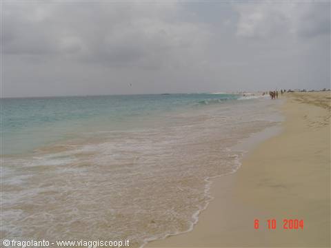 Foamy wave on pale sand, people walking along the shore under cloudy sky