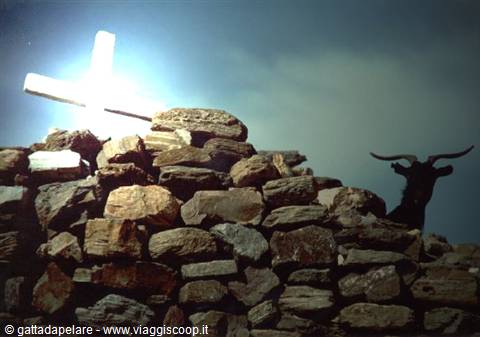 Long-horned goat behind a dry stone wall, white cross against the sky