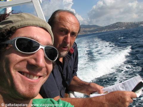 Two men on a ferry, white wake behind and coast with white houses