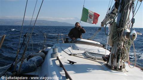 Man steering a sailboat flying the Italian flag, choppy sea, distant coast