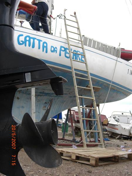 Propeller in foreground, beached white hull on stands, wooden ladder in boatyard
