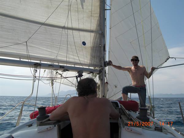 Two men aboard a sailboat, one handling rigging lines at open sea